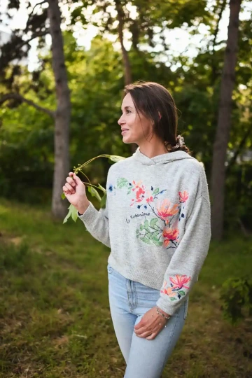 Žena v šedé mikině (fotografka Bára) s květinovou výšivkou drží větvičku a dívá se do strany v přírodním prostředí.