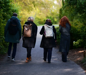 Čtyři osoby kráčející po lesní cestě, zachycené zezadu. Osoba vlevo má na sobě zelený kabát a tmavé kalhoty s batohem, vedle ní jde osoba se světlými vlasy v černém kabátě s hnědým batohem. Další osoba má tmavé oblečení a světlý batoh, zatímco osoba vpravo má výrazné zrzavé vlasy a tmavý kabát. Cestu lemují zelené stromy a keře vytvářející přírodní koridor. Scéna zachycuje společnou procházku v přírodě, pravděpodobně na jaře nebo na podzim.