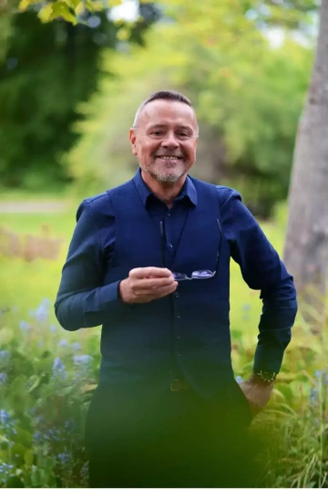 Usmívající se osoba s krátkými světle šedými vlasy a upraveným vousem, stojící v zahradě nebo parku. Má na sobě tmavě modrou košili a sako stejné barvy. V jedné ruce drží brýle a druhou má opřenou v bok. V popředí jsou vidět nízké květiny nebo keře s modrými a fialovými květy. Pozadí tvoří rozostřené stromy a travnatá plocha. Osoba vypadá přátelsky a sebevědomě, s širokým úsměvem do kamery. Na zápěstí má hodinky. Celková atmosféra je příjemná a jarní.