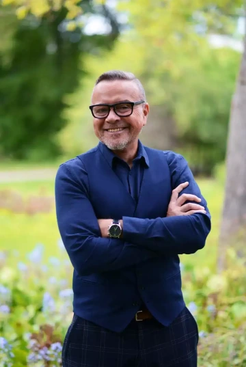 Osoba (Petr Daňko) s brýlemi a krátkými světlými vlasy se usmívá do kamery v parkovém prostředí. Má na sobě tmavě modré sako a košili, na zápěstí má náramkové hodinky s tmavým páskem. Stojí se zkříženýma rukama v sebevědomé póze. V pozadí je rozostřená zeleň stromů a keřů, což vytváří příjemnou přírodní atmosféru. Osoba vypadá profesionálně a přátelsky.