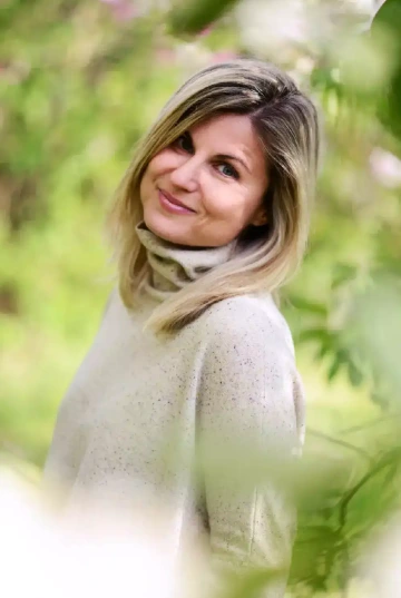 Osoba (Kristýna Tvrdá) s polodlouhými světlými vlasy s tmavšími kořínky, otáčející se přes rameno a usmívající se do kamery. Má na sobě krémový nebo světle béžový svetr s jemným vzorem a světlý šátek nebo rolák kolem krku. Pozadí tvoří rozostřená jarní zeleň, pravděpodobně park nebo zahrada. Portrét zachycuje osobu z polovičního profilu s příjemným, přátelským výrazem ve tváři na přirozeném, světlém pozadí.