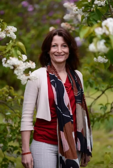 Usmívající se osoba (Lenka Macková) s hnědými vlasy po ramena, stojící mezi kvetoucími keři s bílými květy. Má na sobě červený top, světlé sako s tříčtvrtečními rukávy, světlé kalhoty a barevný šátek s převážně tmavě modrými, červenými a hnědými vzory. Na zápěstí má náramek. V pozadí je rozostřená zahrada nebo park s různými květinami. Osoba vypadá přátelsky a je zachycena v jarním prostředí mezi rozkvetlými keři, pravděpodobně šeříky.