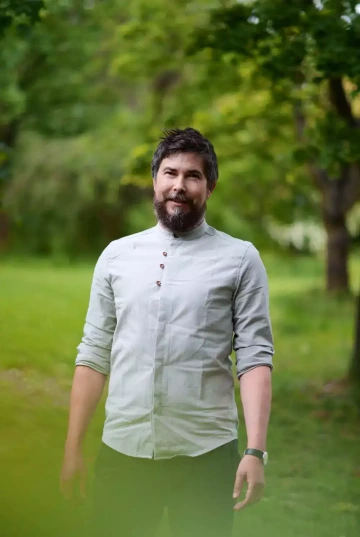 Osoba s tmavými vlasy a vousy stojící na travnaté ploše v parku nebo zahradě. Má na sobě světle šedou košili se stojáčkem a několika knoflíky a rukávy vyhrnutými k loktům. Na zápěstí má hodinky. Dívá se mírně do strany s lehkým úsměvem ve tváři. Pozadí tvoří rozostřené zelené stromy a travnatá plocha, což vytváří příjemné přírodní prostředí. Osoba je zachycena od pasu nahoru v neformálním, ale upraveném vzhledu.