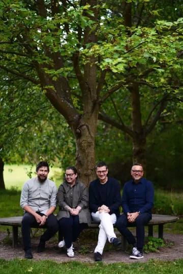 Čtyři osoby (tým Silene) sedící na kulaté lavičce pod stromem v parku. Zleva doprava: osoba s vousy ve světle šedé košili a tmavých kalhotách, osoba s brýlemi v šedé košili a tmavých kalhotách, osoba v tmavě modrém saku a bílých kalhotách s překříženýma nohama, a osoba s brýlemi v tmavě modré košili a tmavých kalhotách. Všichni se usmívají do kamery a sedí na dřevěné kruhové lavičce kolem stromu. Pozadí tvoří zeleň parku nebo zahrady s velkým listnatým stromem nad nimi. Scéna působí přátelsky a uvolněně v přírodním prostředí.