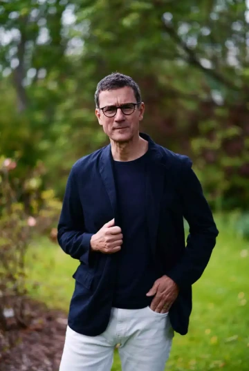 Osoba (Tomáš Zykán) s brýlemi a krátkými tmavými vlasy stojící v parku. Má na sobě tmavě modré sako, tmavý top a světlé kalhoty. Jednou rukou se drží za klopu saka, druhou má v kapse kalhot. Výraz tváře je vážný a soustředěný. V pozadí je rozostřená zeleň stromů a keřů, která vytváří přírodní kulisu. Osoba je zachycena v plné výšce v elegantním, ale neformálním oblečení vhodném pro venkovní prostředí.