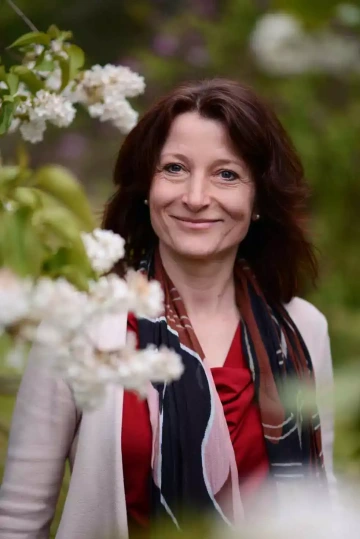 Lenka, členka týmu společnosti Silene, s hnědými vlasy po ramena, usmívající se na portrétu zachyceném mezi kvetoucími bílými květy. Má na sobě červený top, světlé sako a vícebarevný šátek v kombinaci černé, červené a krémové barvy. Portrét je focený v jarním přírodním prostředí s rozostřeným zeleným pozadím. Profesionální fotografie vystihuje přátelský a přístupný výraz tváře.