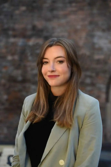 Portrét ženy s hnědými vlasy (Petra Císařová), v černém topu a světle zeleném saku, pózující před cihlovou zdí.