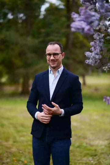 Osoba s brýlemi v tmavě modrém saku a bílé košili, stojící v parku. V pozadí jsou vidět fialové květy, pravděpodobně šeříky. Profesionální portrét člena týmu Silene, Jakuba, v jarním prostředí.