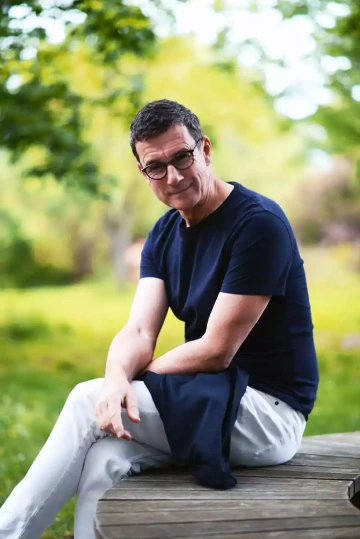 Osoba v tmavě modrém tričku a světlých kalhotách sedící na dřevěné lavici nebo molu v zeleném parkovém prostředí. Neformální portrét člena týmu Silene, Tomáše, v přírodě.