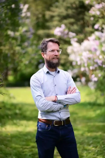 Osoba s vousy a světlou košilí stojící se zkříženýma rukama v parkovém prostředí s kvetoucími keři. Profesionální portrét člena týmu Silene, Václava, v přírodním prostředí.