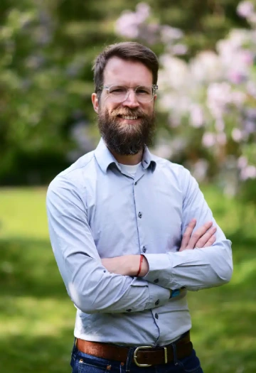 Usmívající se osoba (Václav Jiránek) s vousy a brýlemi s průhlednými obroučkami stojící v parku nebo zahradě. Má na sobě světle modrou košili s bílým tričkem pod ní a na zápěstí má červený náramek. Osoba stojí se zkříženýma rukama v sebevědomé póze a široce se usmívá do kamery. V pozadí jsou rozostřené kvetoucí keře a stromy s náznakem fialových nebo růžových květů, pravděpodobně šeříky. Celkový dojem je přátelský a uvolněný, zachycený v jarním prostředí.