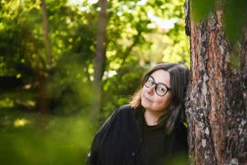 Tmavovlasá žena (Zuzka, zakladatelka Silene) v brýlích opřená hlavou o strom, dívající se do záběru.