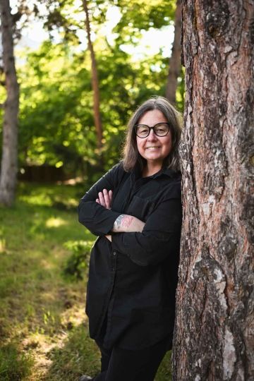 Tmavovlasá žena Zuzka (zakladatelka Silene) usmívající se a stojící s překříženýma rukama opřená o strom v přírodě.