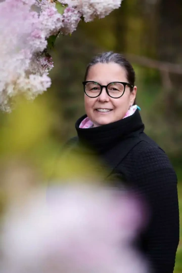 Usmívající se osoba (Zuzana Jiránková) s černými obroučkovými brýlemi a vlasy staženými dozadu, zachycená pod rozkvetlým šeříkem s jemně růžovými květy. Má na sobě černý svetr nebo kabát s vysokým límcem a pod ním je vidět růžový nebo barevný šátek. Osoba se širokým úsměvem hledí do kamery. V popředí a kolem jsou jemně rozostřené květy šeříku a zelené listy, vytvářející měkký, jarní rámec. Fotografie využívá mělkou hloubku ostrosti, díky které vyniká obličej osoby na rozostřeném pozadí.